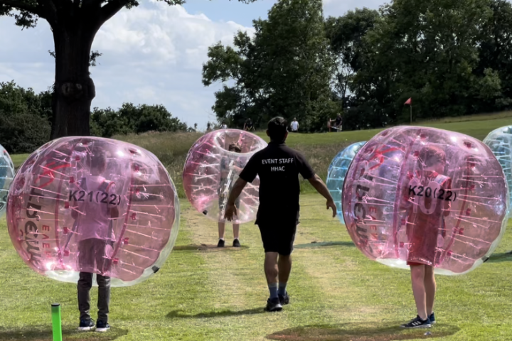 People in pink bubble balls on grass with trees and event staff member.