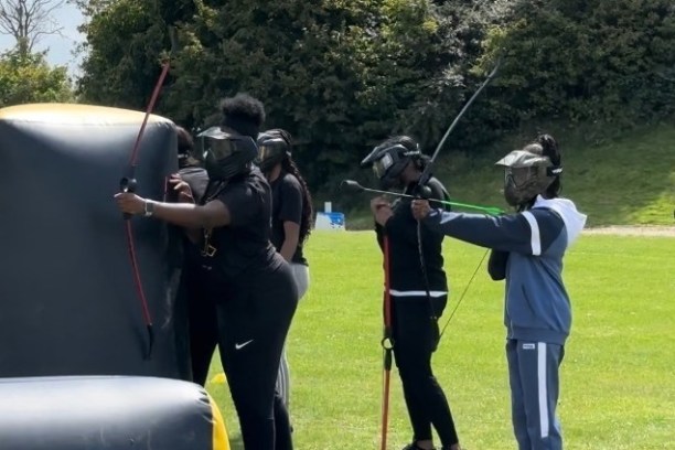 People with masks holding bows and aiming at targets in an outdoor field.