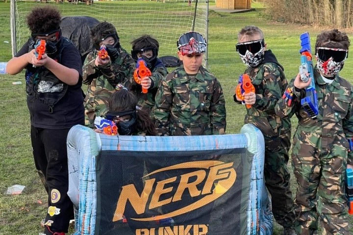 Children in camo outfits aiming Nerf guns behind a barrier outdoors.