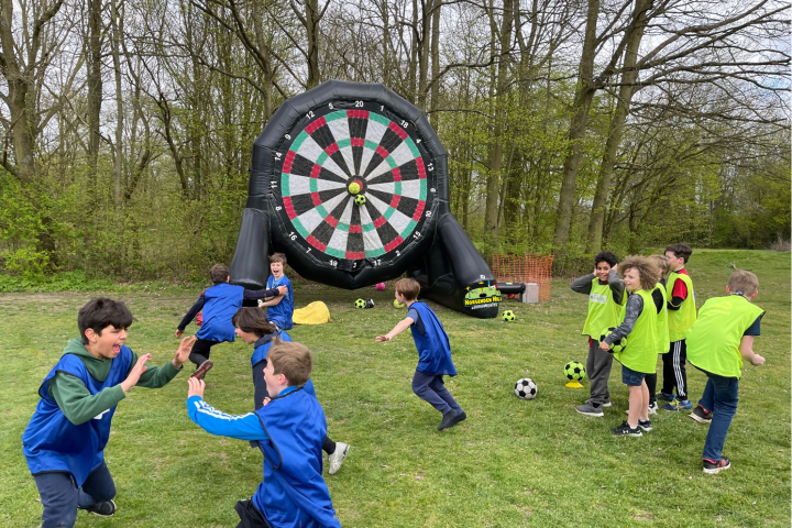 Children playing soccer in front of a large inflatable dartboard outdoors.