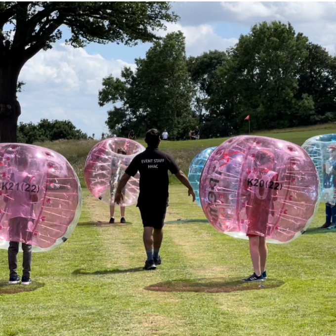 People in inflatable bubbles playing in a grassy park under a cloudy sky.