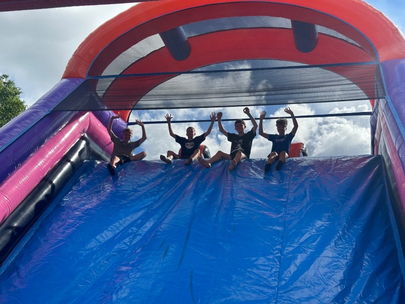 Four people sitting atop a large inflatable slide, hands up, beneath a cloudy sky.