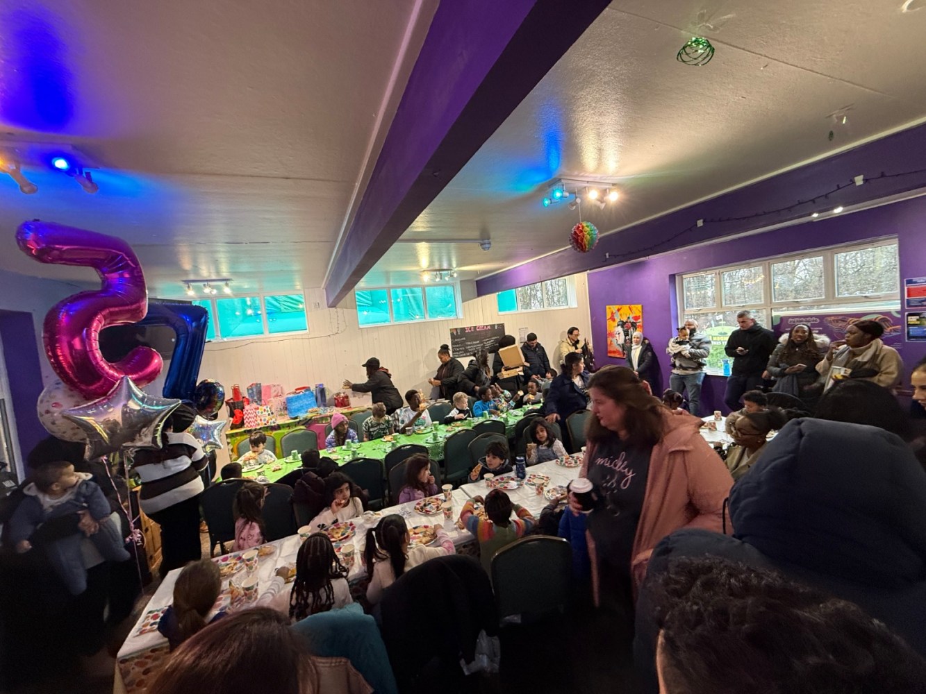 Crowded birthday party with people eating and balloons in a colorful room.