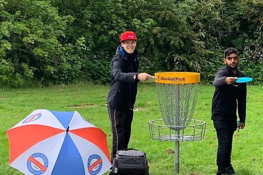 Two people playing disc golf beside a basket, with an umbrella and bag on the grass.