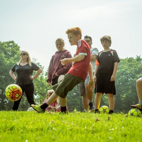 Children playing soccer on a grassy field with trees in the background.