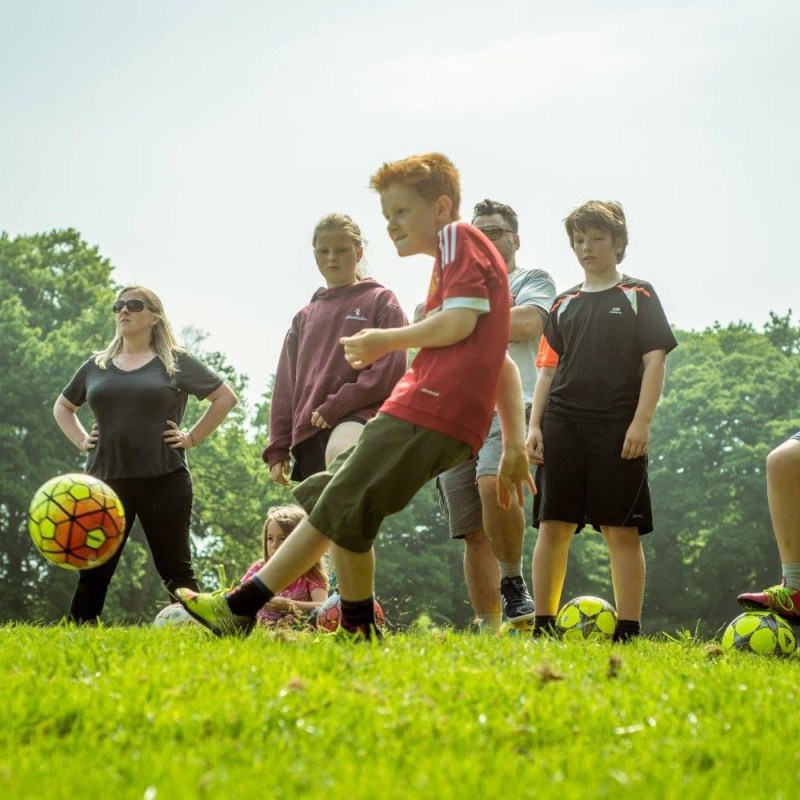 Children playing soccer on a grassy field with trees in the background.