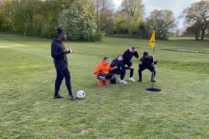 Four people crouching by a golf flag while one stands with a soccer ball on a grassy field.