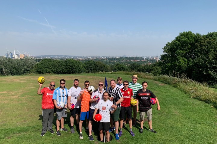 Group of men in soccer jerseys holding balls on a grassy hill with city skyline in background.