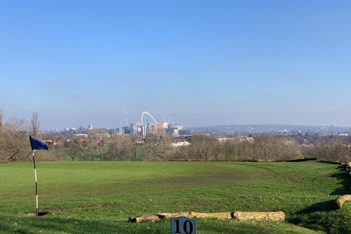 Golf flag on a grassy hill with city skyline and stadium arch in background under clear blue sky.