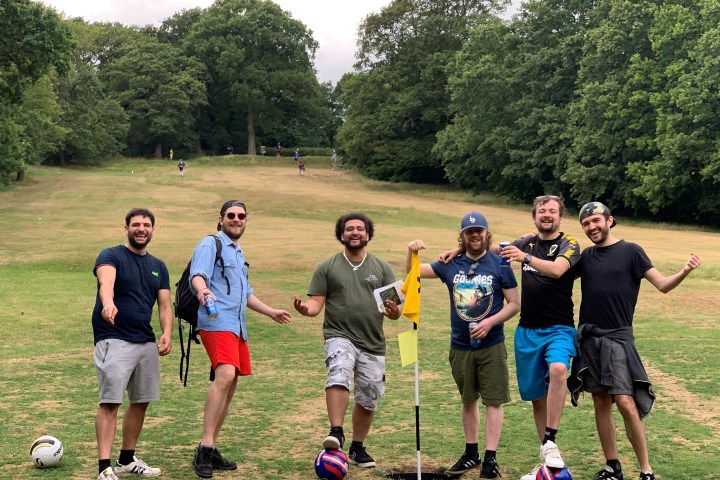 Six men smiling on a grassy field, near a flag and soccer balls, with trees in the background.