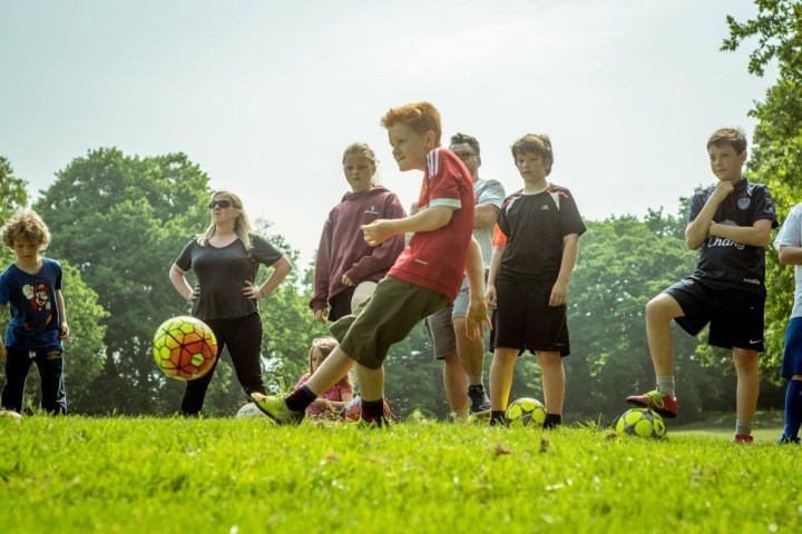 Children playing soccer in a grassy park, one kicking a ball while others watch.