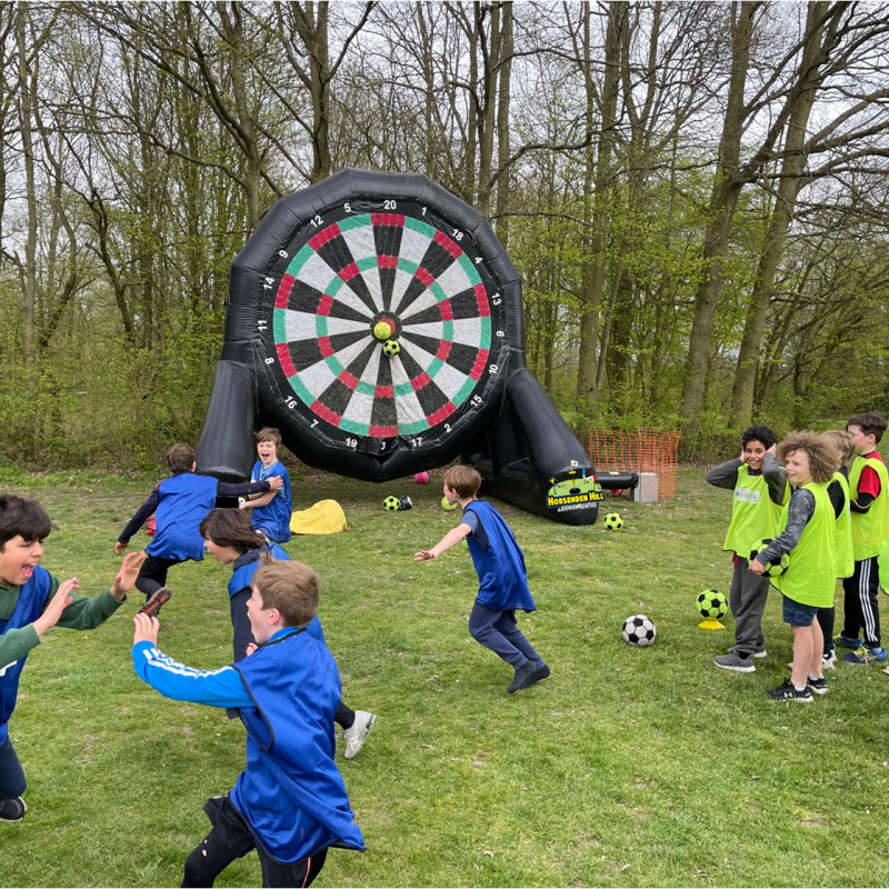 Children in blue and green vests playing soccer near a giant inflatable dartboard outdoors.