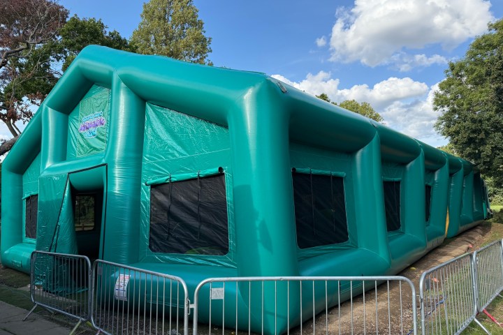 Large green inflatable structure with windows, fenced off in a park on a sunny day.