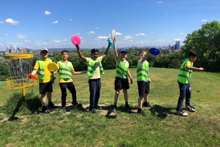 Six children in vests holding frisbees on a grassy hill near a disc golf basket.