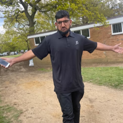 Person with outstretched arms holds a phone, standing on a path with trees and building in background.
