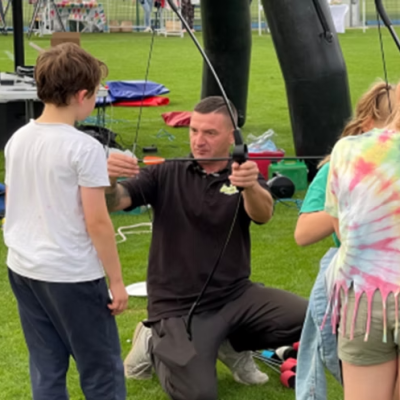 Man kneeling and talking to two children on a grassy field with equipment in the background.