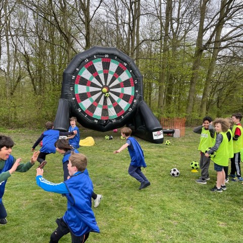 Children playing soccer near a giant inflatable dartboard outdoors.